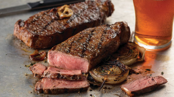 Two grilled New York strip steaks, one sliced showing medium-rare doneness. Served with an amber beer.