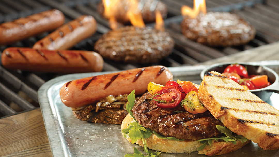 cooked burger with tomatoes and lettuce and hot dog on silver tray with grill in background with burgers and hot dogs