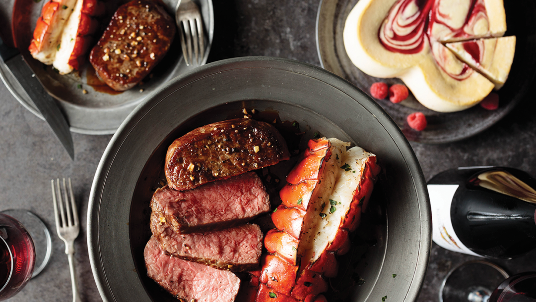 Medium-rare cooked Butcher's Cut Ribeye, Lobster Tail, red wine, and heart shaped cheesecake for a romantic Valentine's Day meal at home
