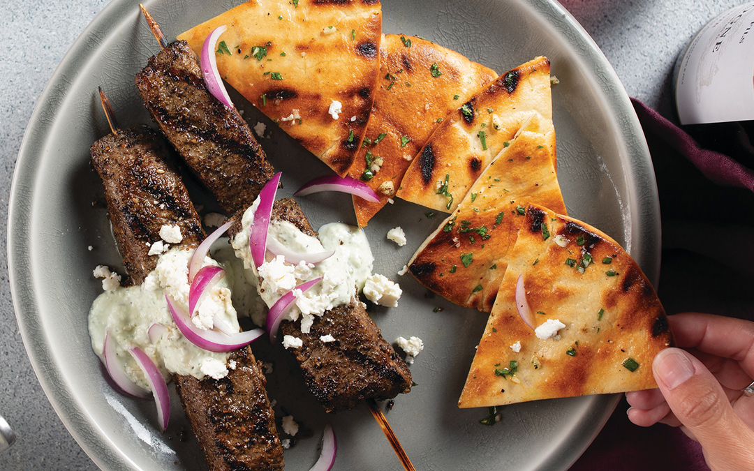 Two Mediterranean-style grilled Filet Mignon steak kabobs on bamboo skewers covered with tzatziki sauce and sliced red onions and served with grilled seasoned pita bread