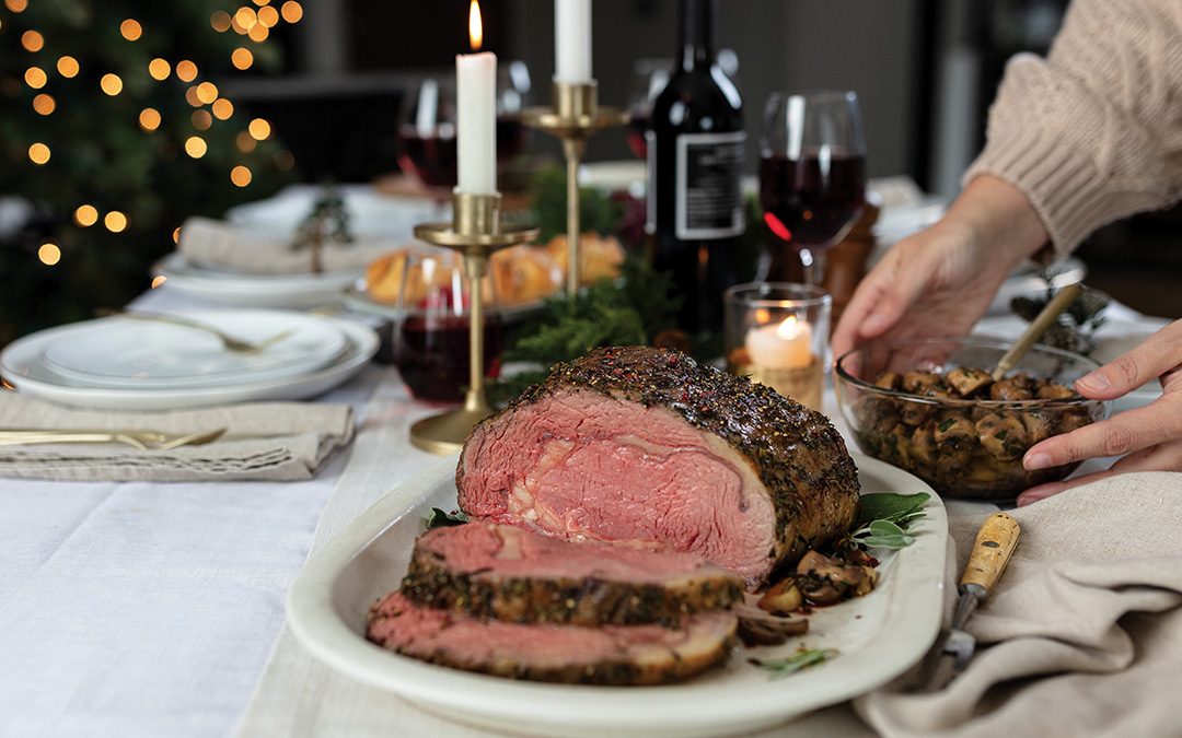 Dijon-Herb Prime Rib Roast sliced on platter and served with garlic butter mushrooms and red wine on a table set for a holiday gathering