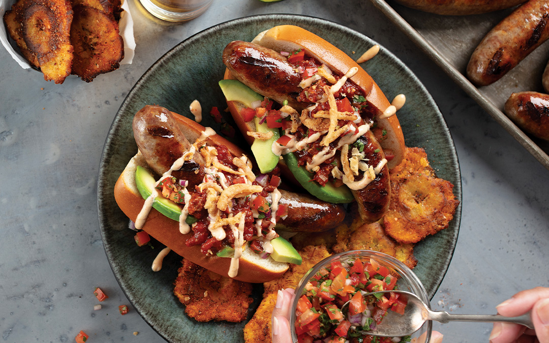 2 Mexican Bratwurst Dogs with grilled bratwurst in a toasted bun topped with chorizo chili, avocado slices, pico de gallo, chipotle aioli, and crispy fried shallots. Served on a silver platter with BBQ Tostones.