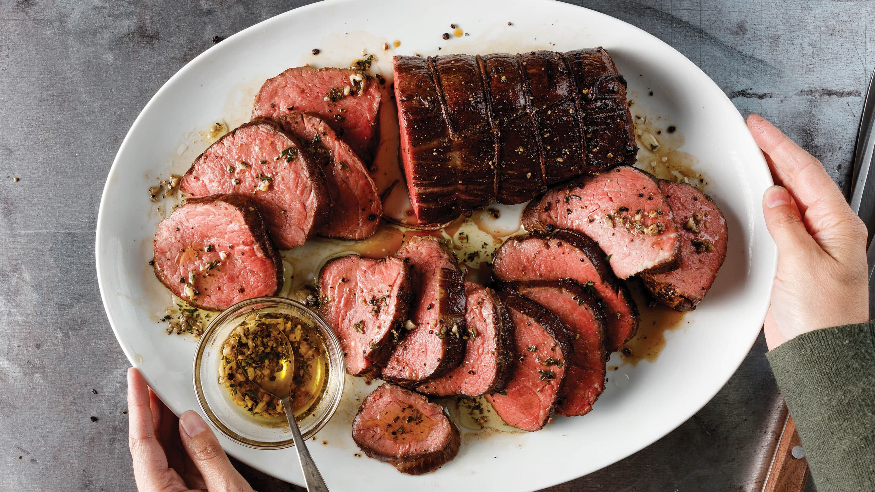 How to sear roast chateaubriand roast Person holding white platter with sear-roasted chateaubriand cooked medium-rare and sliced. Served with a small bowl of garlic and herb melted butter sauce.