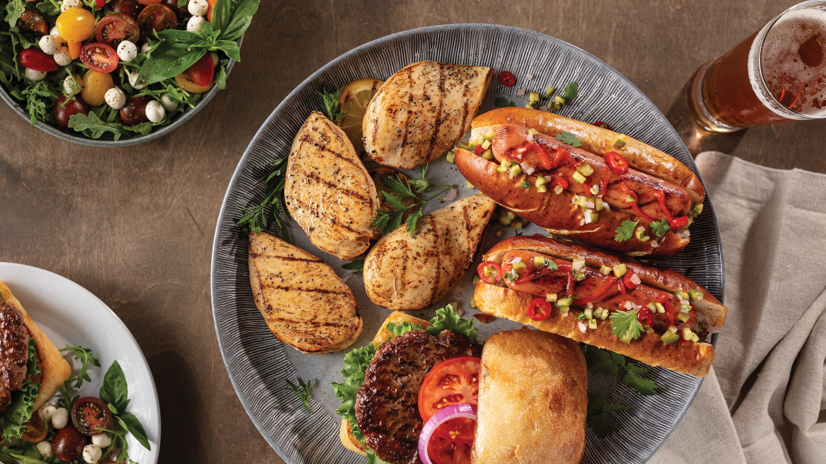how to fight shrinkflation at the grocery store with Omaha Steaks Large serving platter with 4 chicken breasts, 2 gourmet franks with toppings, burger with lettuce, tomato and red onion. Serving bowl with side salad. Smaller plate with burger and salad. Served with glass of water and glass of beer.