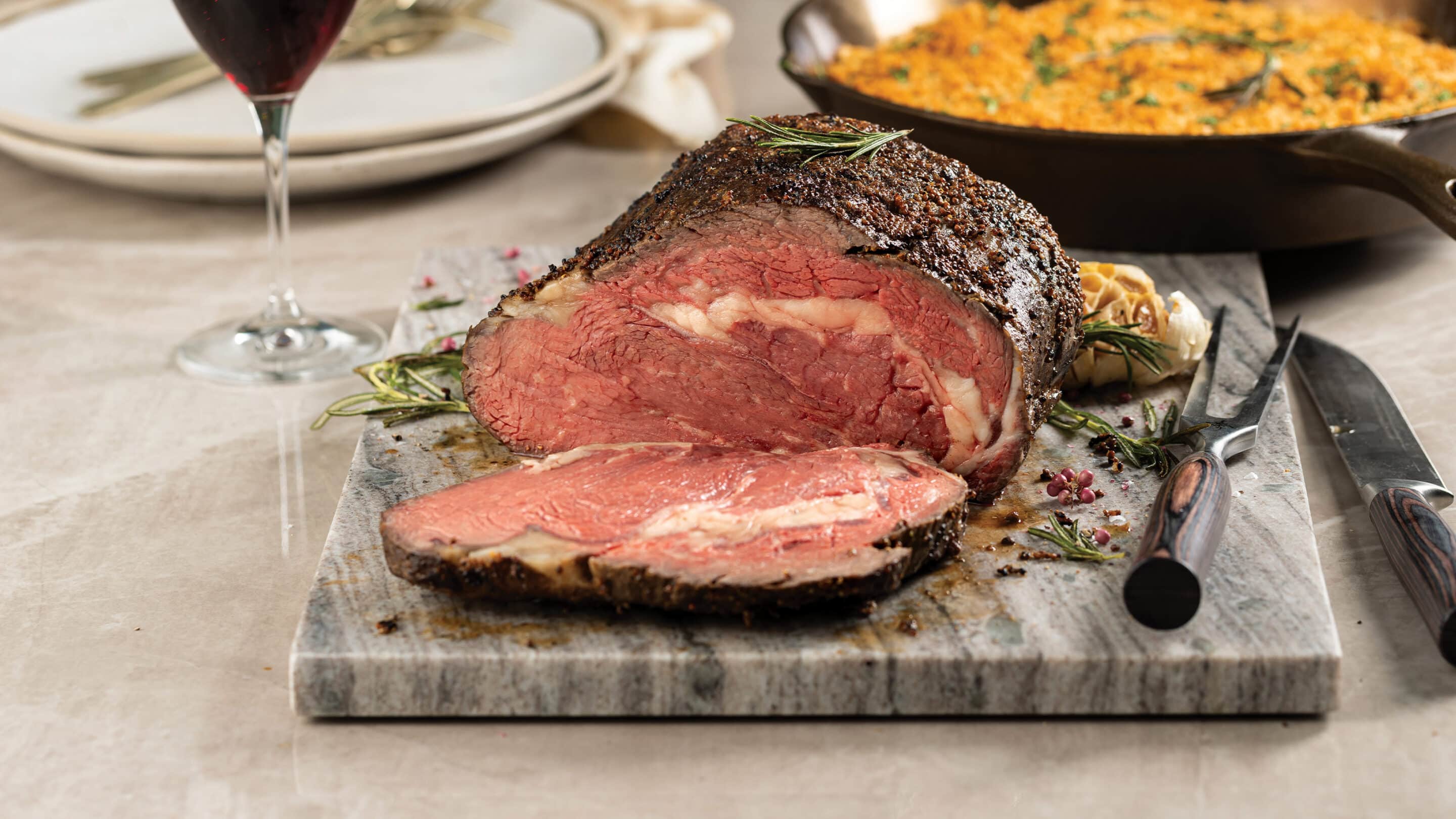 Dijon-rubbed prime rib cooked medium rare and sliced on a marble cutting board. Cast iron skillet with cheddar crumb mashed potatoes, glass of red wine and plates in background.