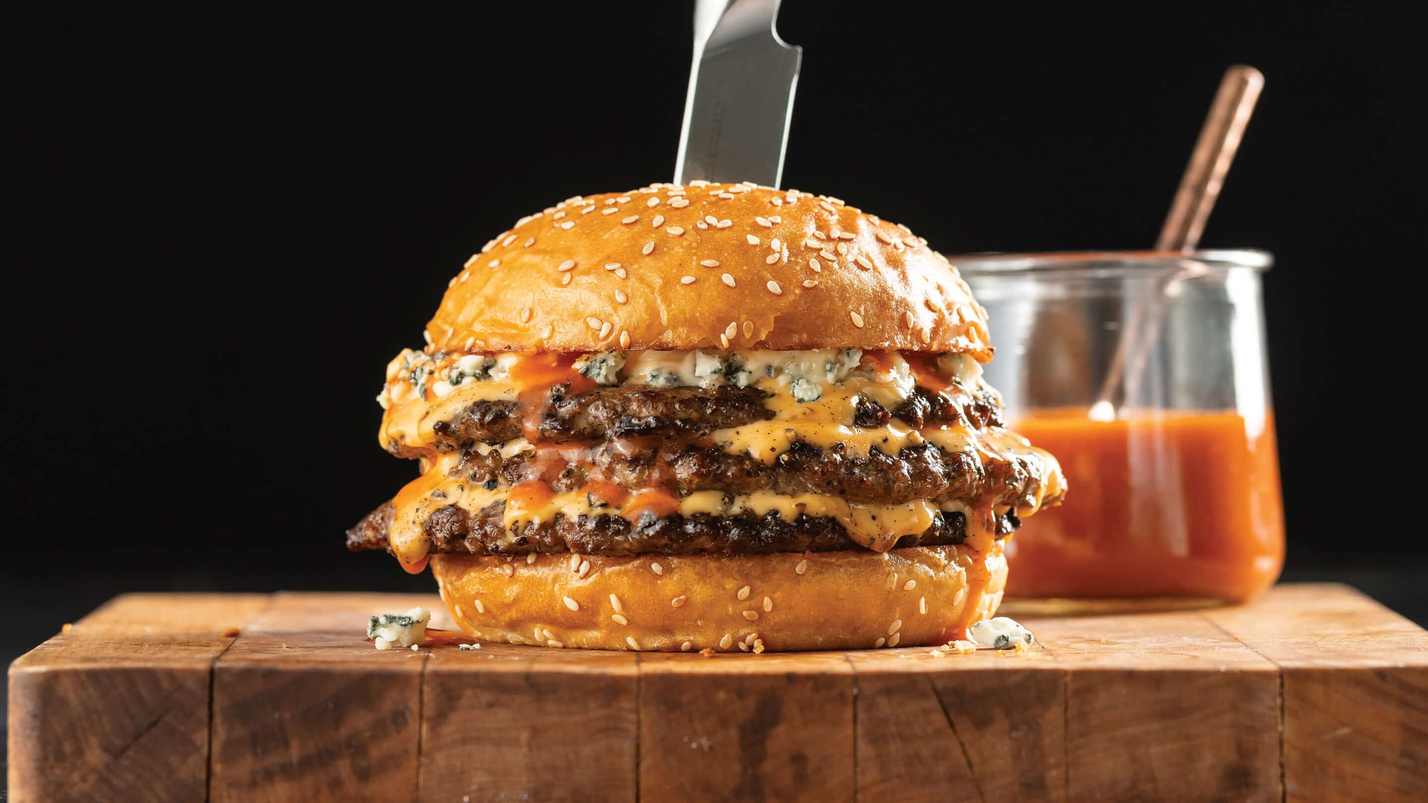 Triple-Decker Buffalo Smash Burger With Blue Cheese Mayo on a wooden cutting board with glass bowl of Buffalo sauce behind it.