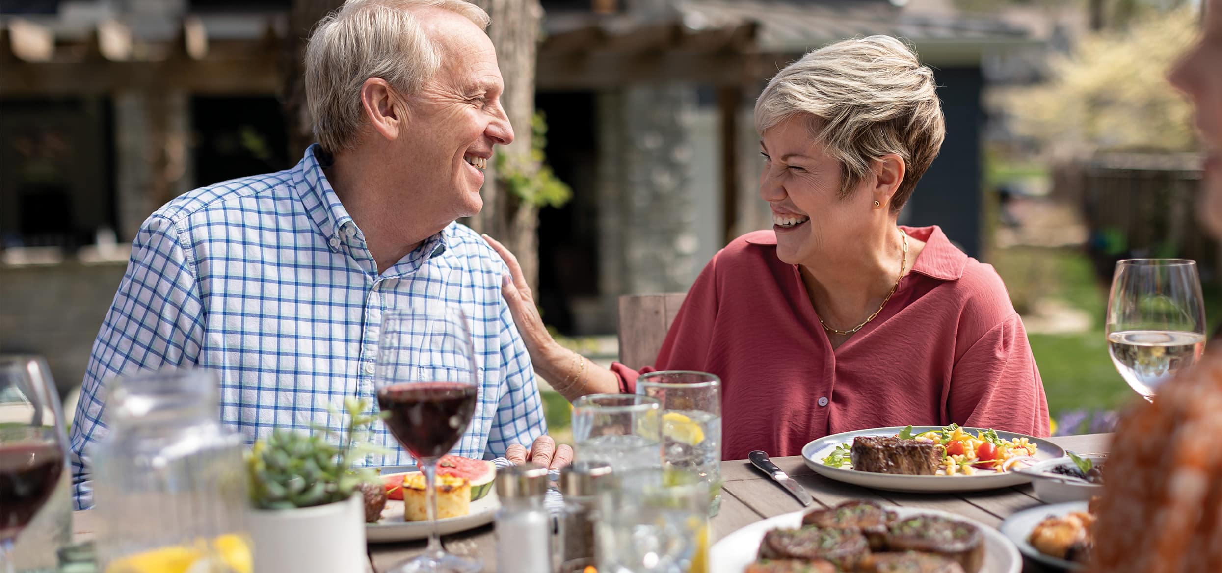 A smiling older couple sits at an outdoor dining table, enjoying a meal together. The woman, wearing a red blouse, laughs while touching the man’s shoulder, and the man, in a light blue checkered shirt, smiles back at her. Plates of food, glasses of water with lemon, and a glass of red wine are on the table, with a backyard setting and house visible in the background.