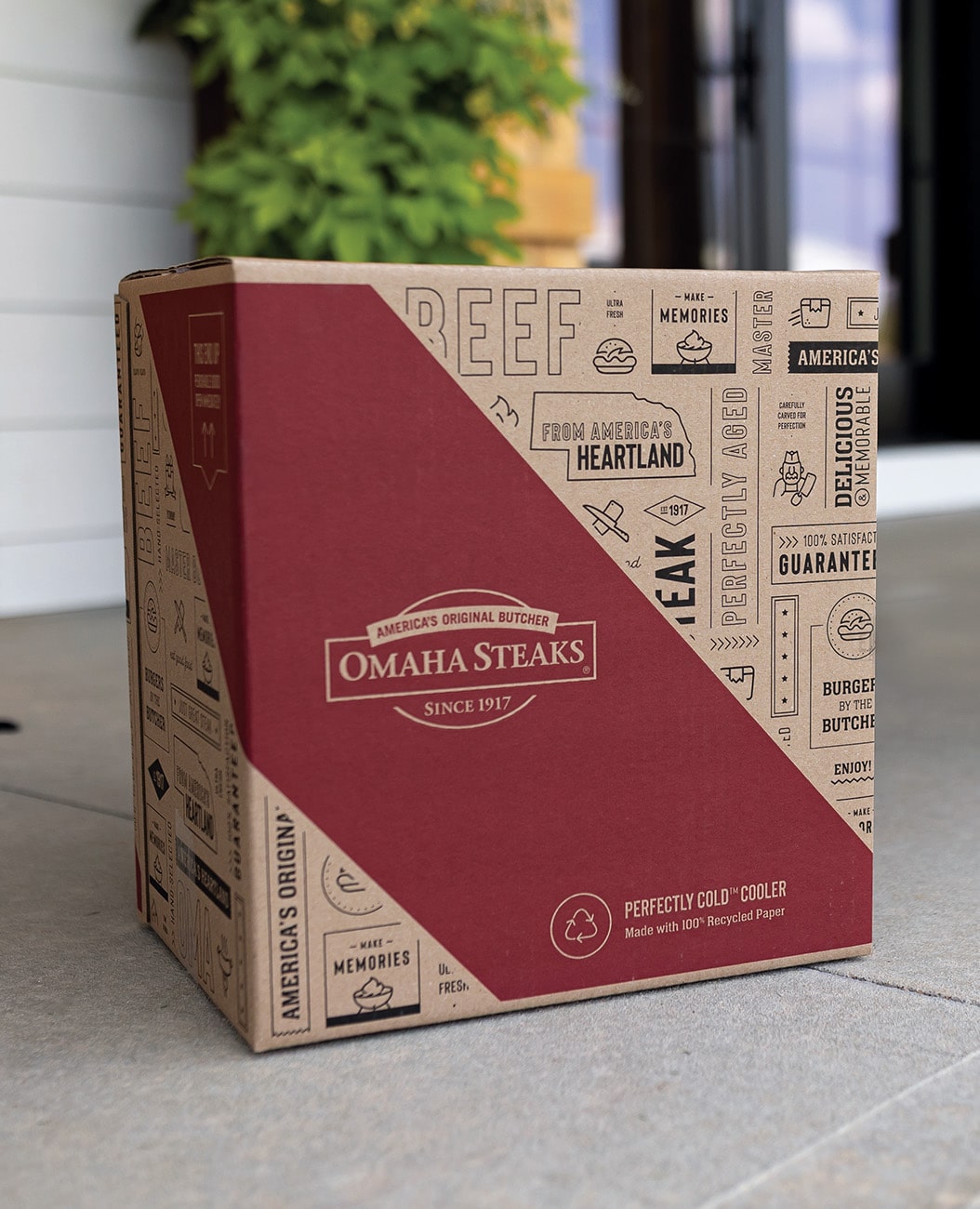 A branded cardboard shipping box from Omaha Steaks sits on a doorstep. The box features a red and tan design with printed illustrations and text such as “America’s Original Butcher,” “Since 1917,” and “From America’s Heartland,” along with icons related to food and grilling. A blurred green plant and doorway are visible in the background.