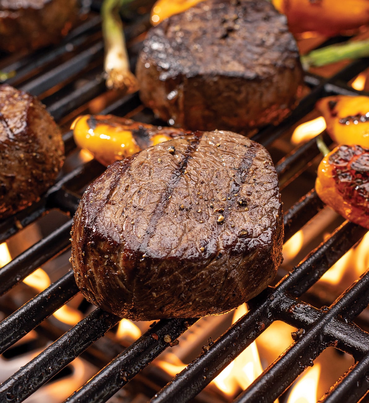 A close-up of thick, round steaks grilling over open flames on a barbecue. The meat has visible grill marks and a seared, seasoned exterior. Around the steaks, vegetables like peppers and green onions are also cooking, with flames flickering beneath the grill grates.